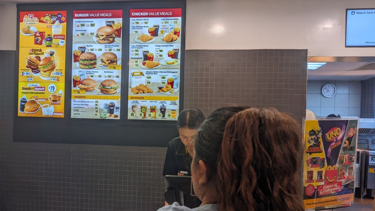 マクドナルド店内の注文カウンター上部に設置されたメニューボード。バーガー類やチキンセットの価格と写真が並び、手前には注文を行う客の後頭部と、レジで対応する店員の姿、UNOのキャンペーン告知が見える