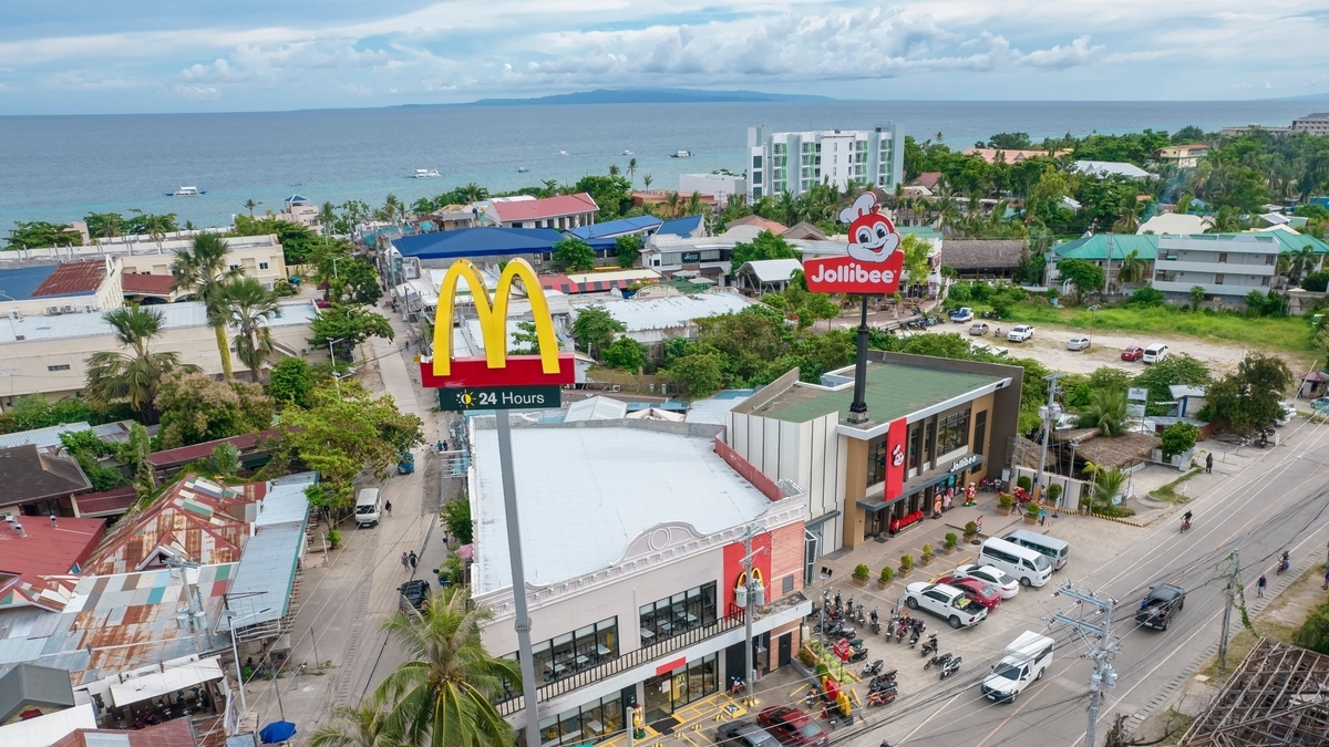 海沿いに位置するマクドナルドとジョリビーの店舗を上空から捉えた鳥瞰画像。手前にはマクドナルドの黄色い「M」の看板、奥にはジョリビーの赤い看板が立ち、周辺には駐車場やヤシの木、遠くに青い海が広がっている
