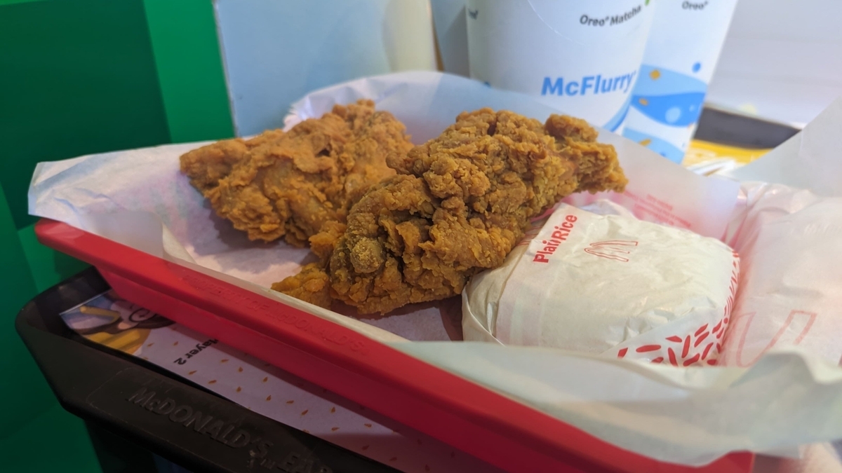 赤いトレイの上に置かれた、マクドナルドのフライドチキン2ピースと、白い紙に包まれた「Plain Rice」のセット。背景には「McFlurry」と書かれた紙コップが並んでおり、店内のトレイ上の様子を接写している