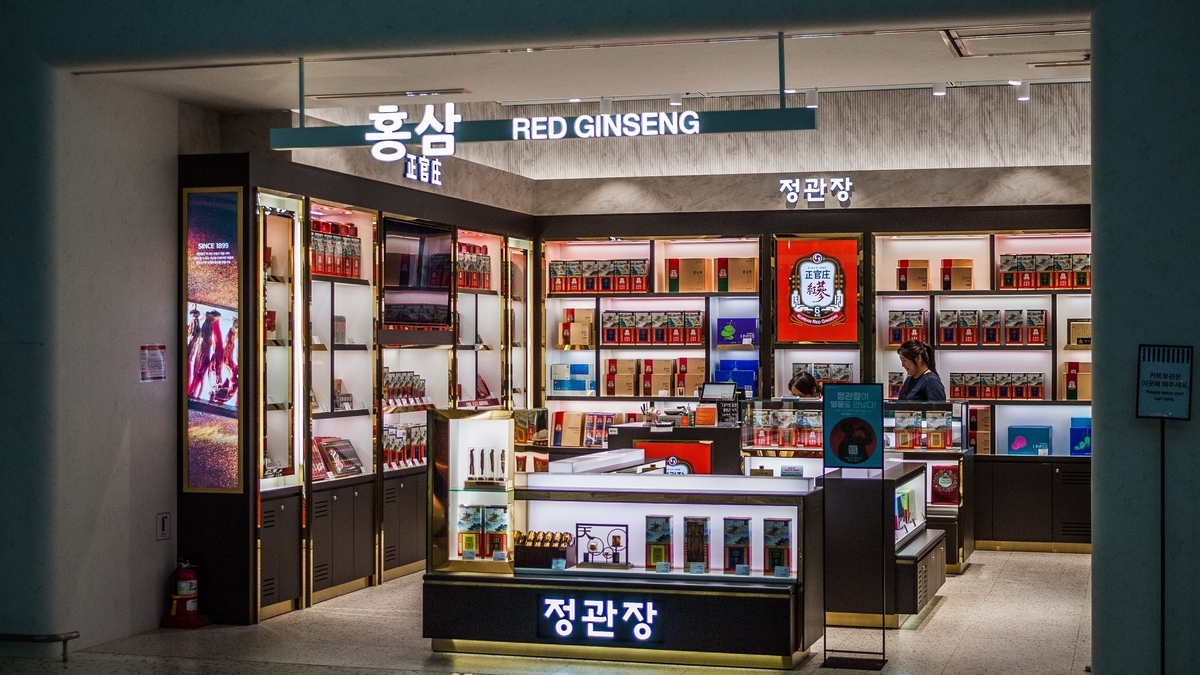 「RED GINSENG(紅参)」の看板を掲げた、韓国の伝統的な健康食品店。木目調の落ち着いた棚には赤いパッケージの商品が整然と並べられ、店員がカウンターで接客を行っています。