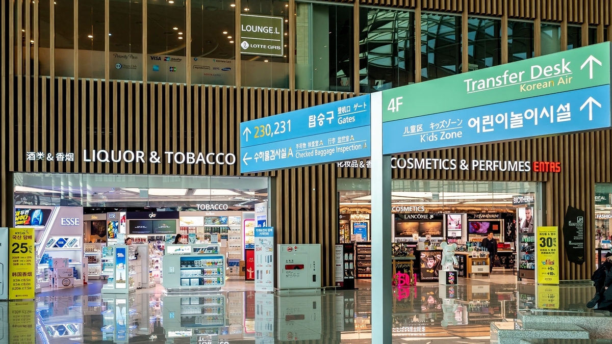 「LIQUOR & TOBACCO」と書かれた空港内の免税店。店舗の前にはゲート案内やキッズゾーンを示す青と緑の大きな案内標識があり、フロアの光沢ある床に店内の明かりが反射しています。