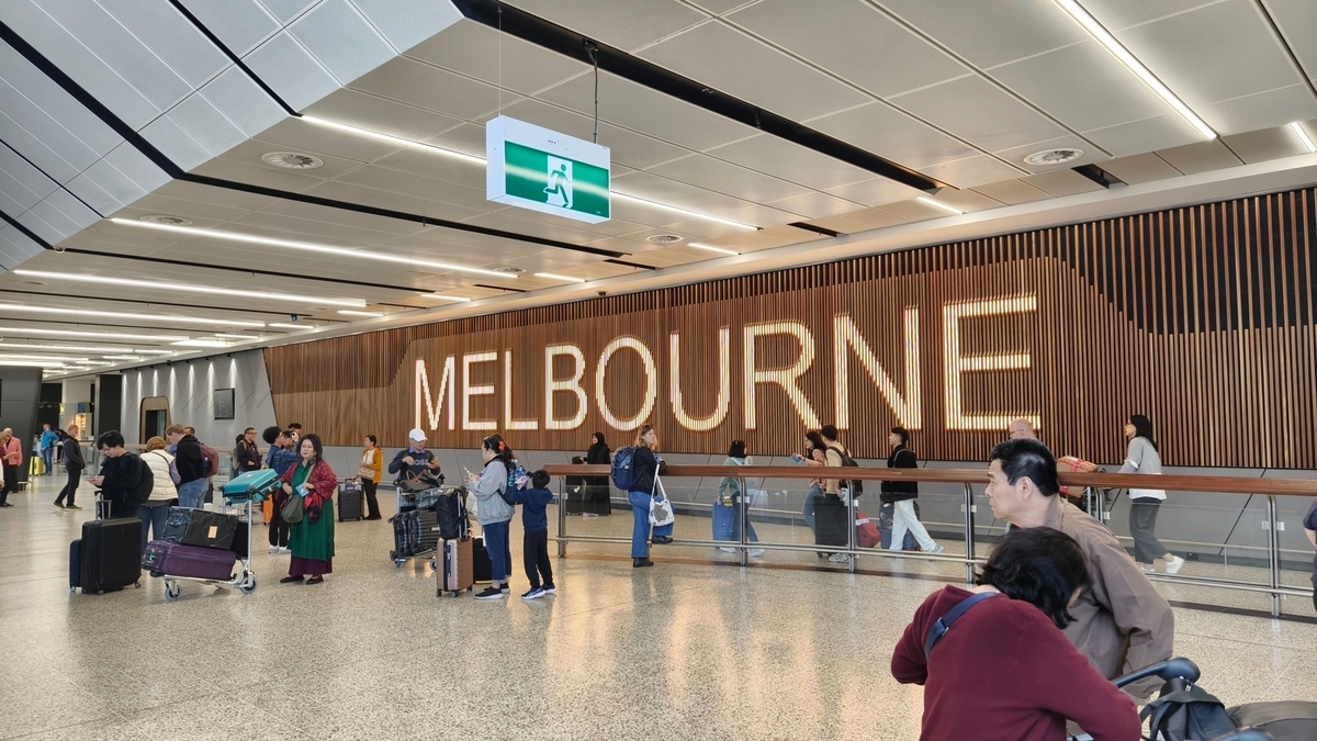 木目調の壁に大きく「MELBOURNE」の文字が光るメルボルン空港のロビー。スーツケースやカートを持った多くの旅行者が行き交い、天井からは緑色の非常口誘導灯が吊り下げられています