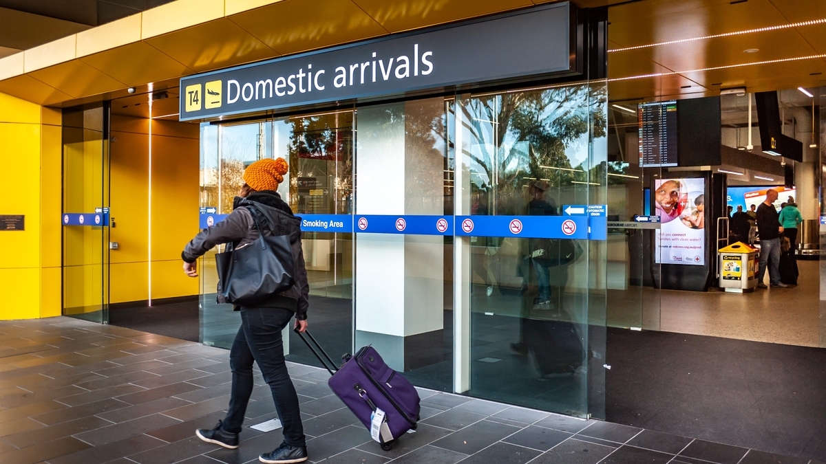 メルボルン空港の第4ターミナル（T4）にある国内線到着口（Domestic arrivals）。黄色い壁が印象的な入り口付近を、紫色のキャリーケースを引いた旅行者が通り過ぎる様子が写っています。