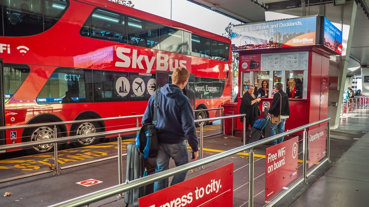 空港のバス乗り場に停車する赤い2階建ての「SkyBus」。手前にはキャリーケースを持つ旅行者が歩いており、右側にはチケット販売窓口と行き先案内が表示されたデジタルサイネージが設置されています