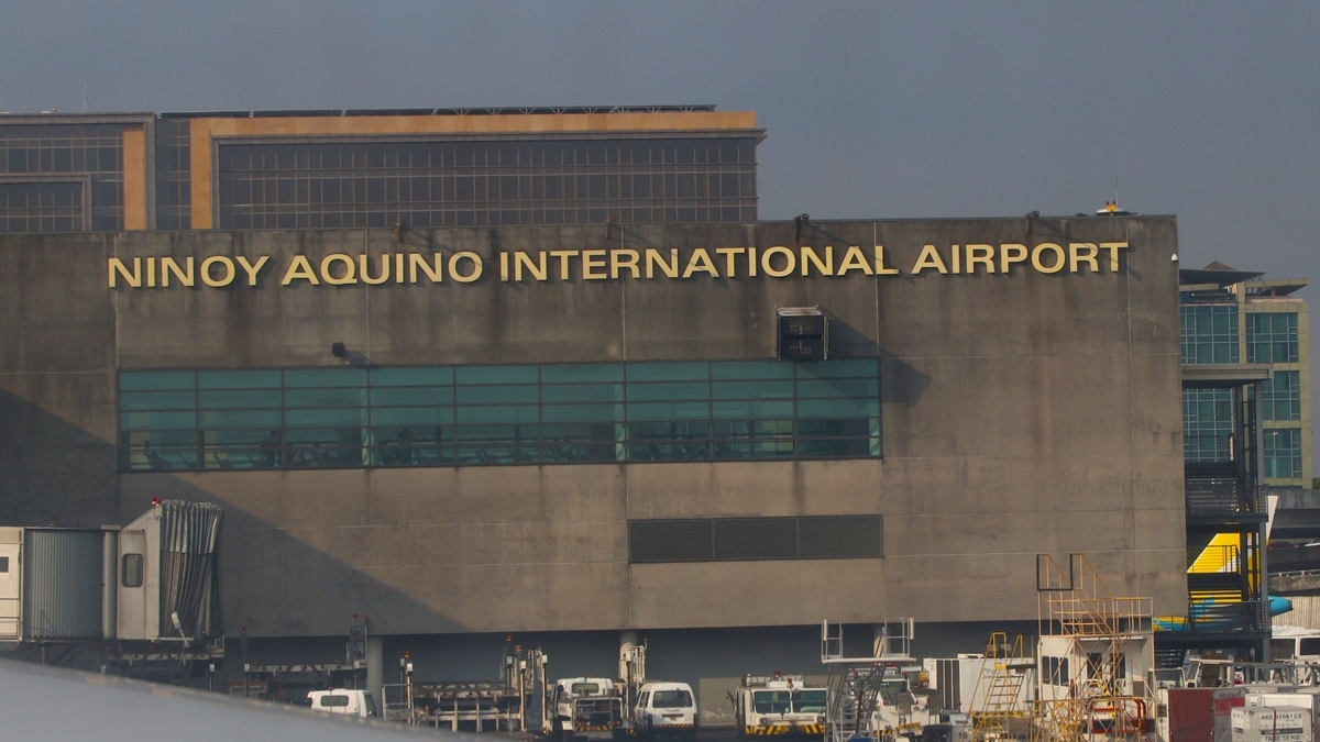 「NINOY AQUINO INTERNATIONAL AIRPORT」の文字が掲げられた空港のターミナルビル。建物の前にはボーディングブリッジが接続され、地上には白い作業車両や階段などの空港機材が配置されています