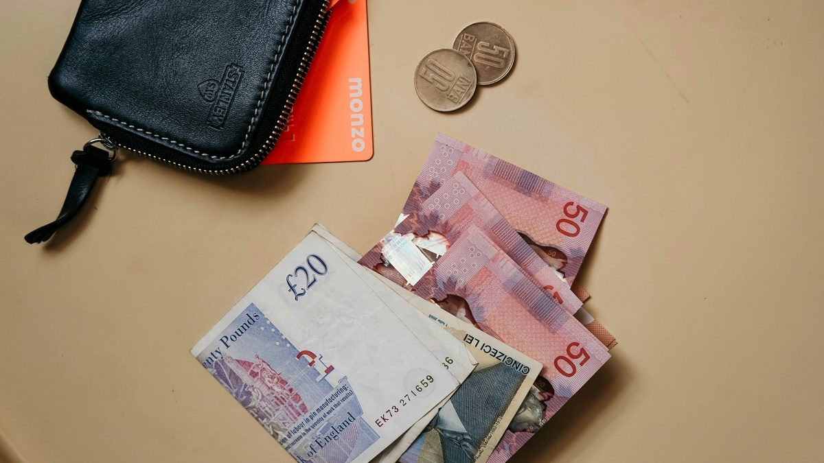 ベージュのテーブルの上に置かれた黒い革財布と紙幣。イングランド銀行発行の20ポンド紙幣、ルーマニアの50レウ紙幣、オレンジ色のMonzoカード、そして2枚の硬貨が散らばっている