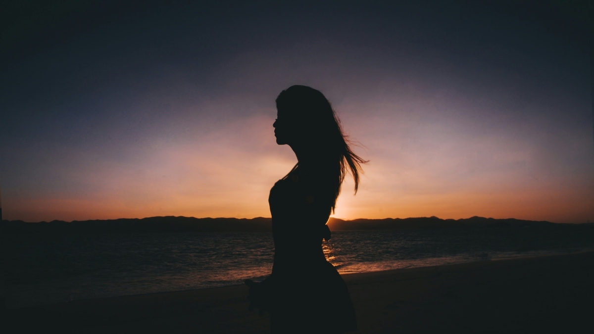 オレンジ色に染まる夕暮れの海辺で、長い髪を風になびかせながら横を向いて立つ女性のシルエット