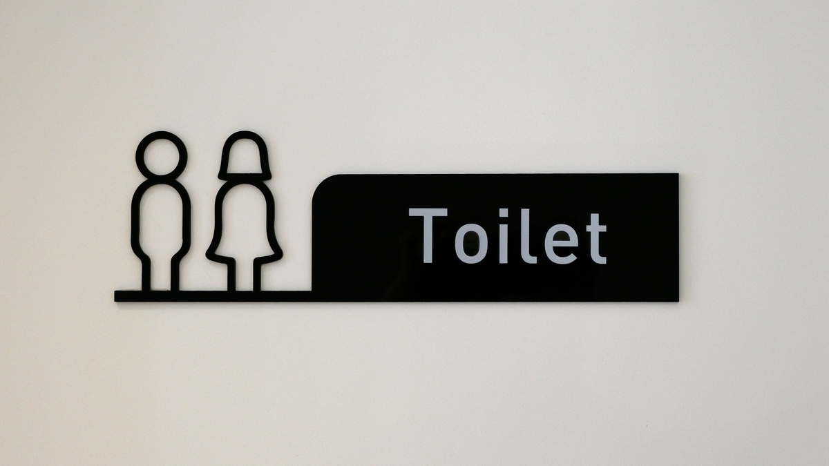 白い壁に取り付けられた、男女のシルエットのピクトグラムと「Toilet」という文字が書かれた黒いトイレの案内看板