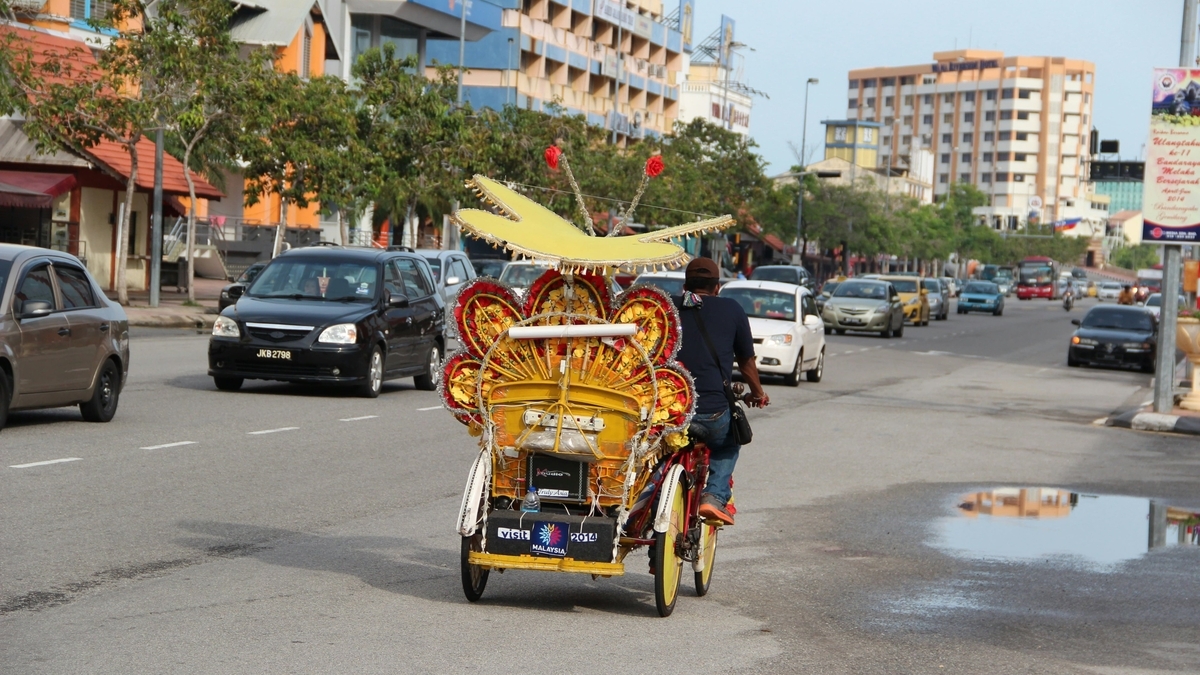マレーシアの街中を走る、黄色い装飾と大きな蝶のような飾りが施された三輪車のリクシャーと、それを運転する男性の後ろ姿