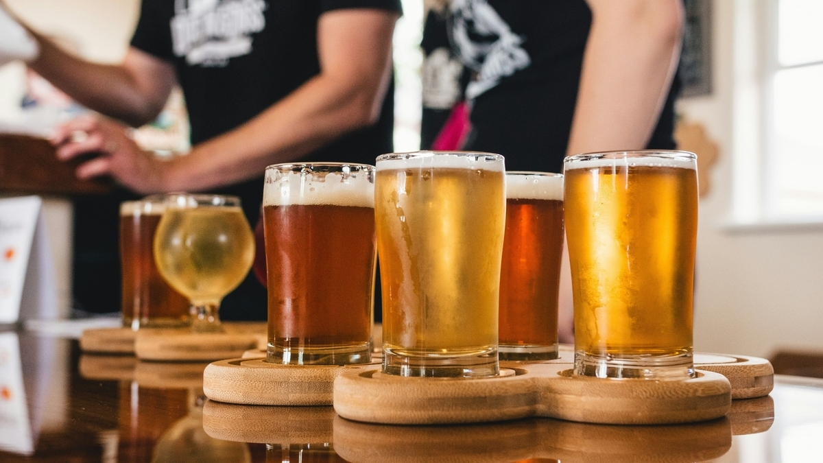 木のカウンターに並べられたテイスティング用のビール。透明なグラスに注がれた黄金色や琥珀色のビールが、専用の木製トレイに5杯セットされ、背景には店員の姿が見えます。