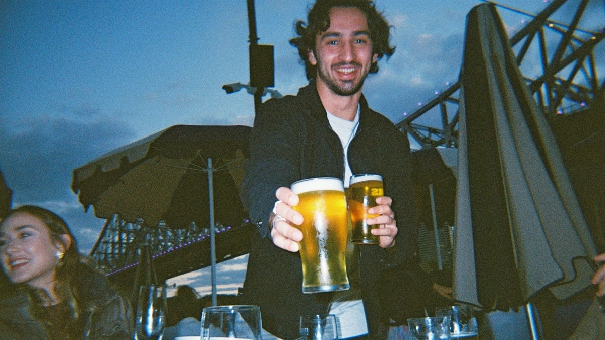 夜の屋外テラスで、黒い上着を着た男性が満たされた2つのビールグラスをこちらへ差し出し、笑顔を見せている様子。背景にはライトアップされた橋の構造物が見えます
