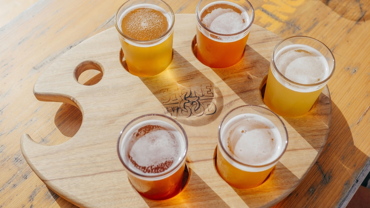 Stone & Woodのロゴが刻印された魚のような形の木製パドルに、5種類の異なる色合いのビールがグラスに入って並んでいる様子。明るい日差しが差し込むテーブルの上で撮影されています。