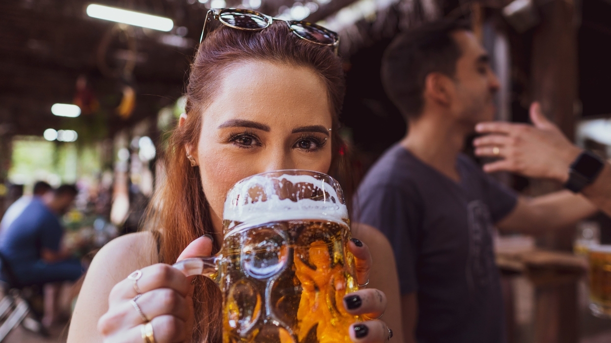 サングラスを頭にのせた女性が、大きなジョッキに注がれた泡立ちの良いビールを両手で持ち、口元に運んで飲んでいる様子。背景にはぼけた人物とパブのような店内が写っています