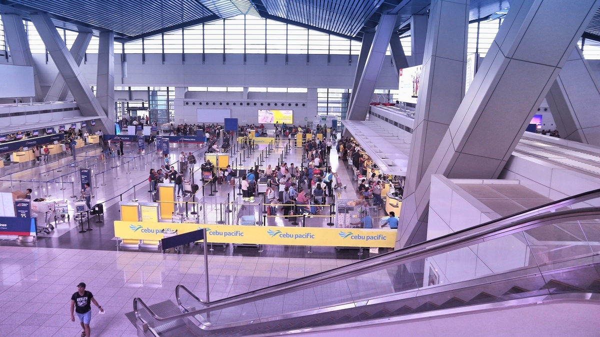 吹き抜けがある広い空港のチェックインカウンターの様子。黄色いCebu Pacificのパーテーションに沿って多くの利用客が並び、手前には下りエスカレーターの側面が写っている