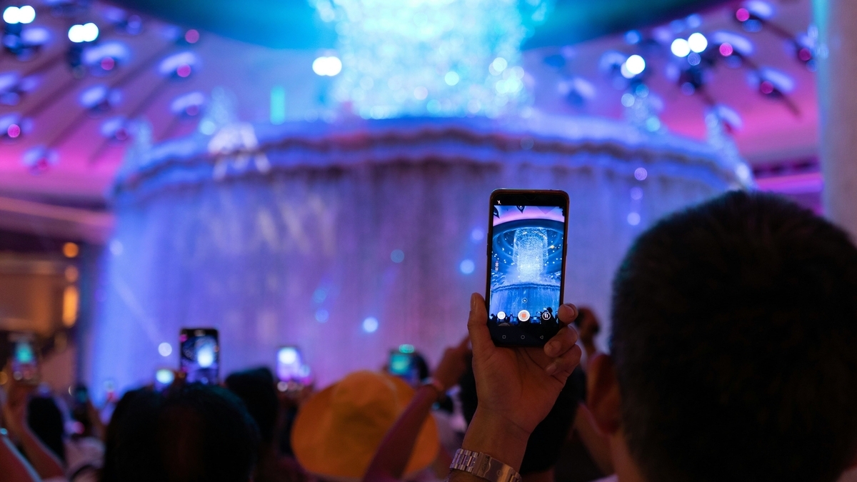 青や紫の照明に照らされた華やかな屋内イベント会場で、多くの観客がスマートフォンを掲げて正面の巨大な噴水を撮影している様子
