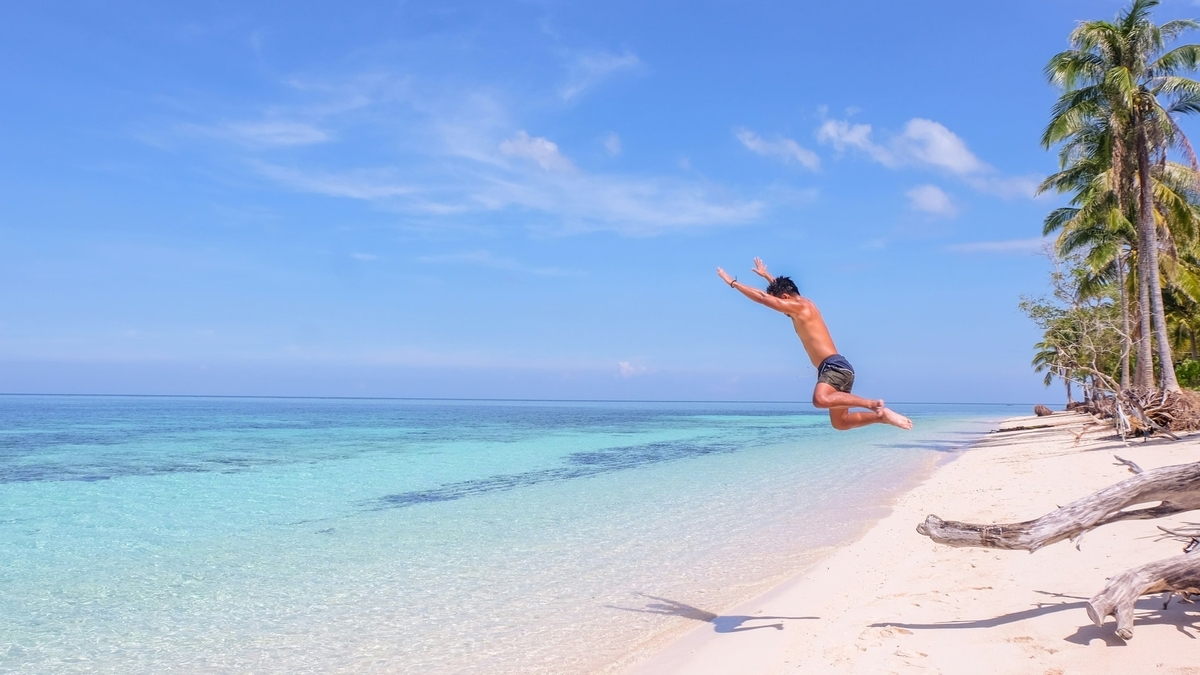 どこまでも続く青い空と透明度の高い海が広がる白い砂浜で、上半身裸の男性が波打ち際に向かって勢いよくダイブしている躍動感のある瞬間