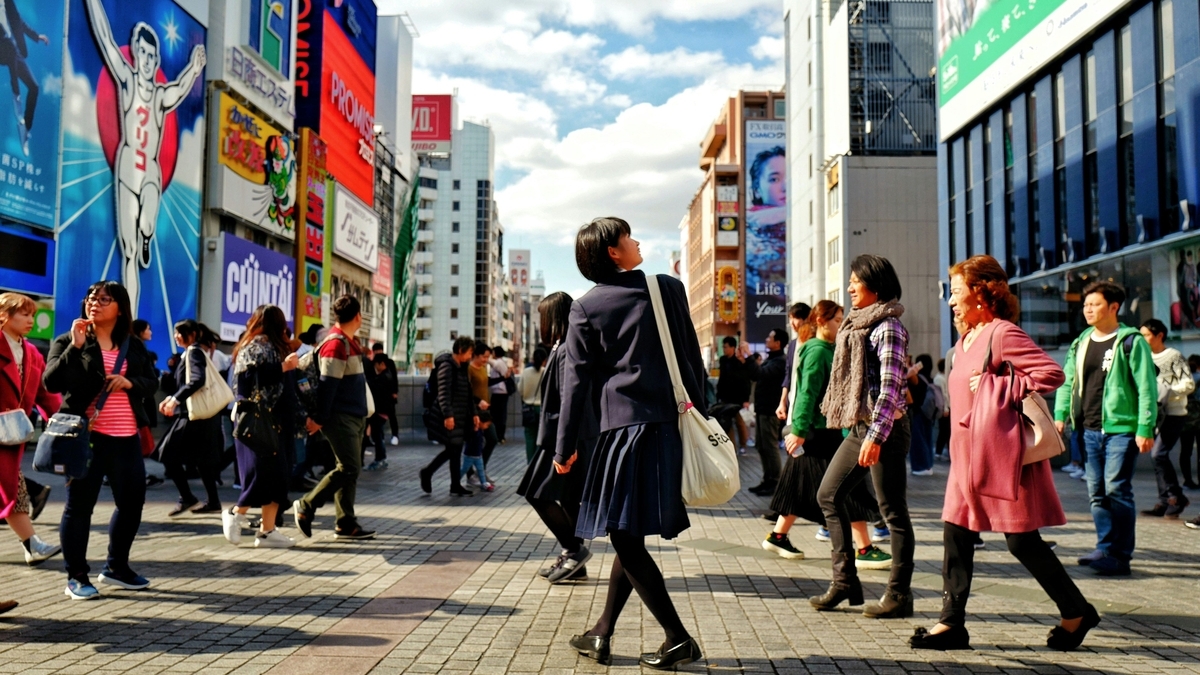 賑やかな都市の広場を多くの人々が行き交っている。手前には制服姿の生徒やピンク色の上着を着た女性が歩いており、背景のビルには多くの看板や広告が掲げられている