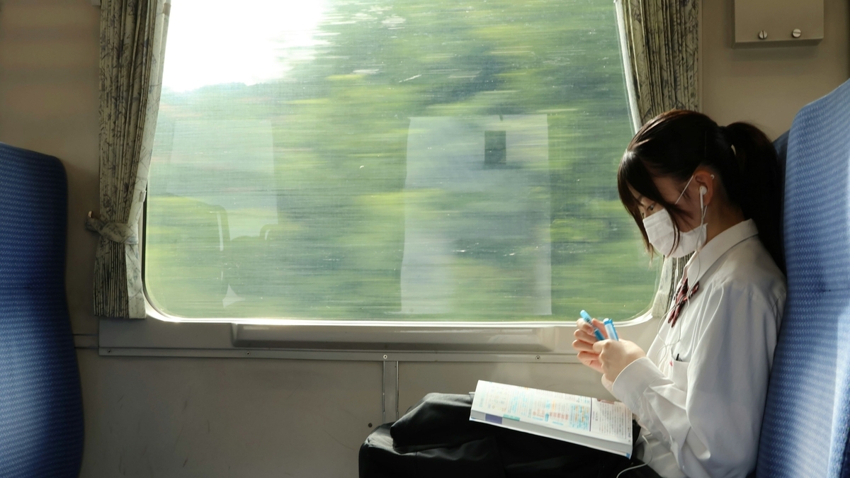 列車の座席に座った女性が本を広げ、手には2本の青いペンを持っている。女性はマスクとイヤホンをしており、窓の外には緑色の風景が流れている