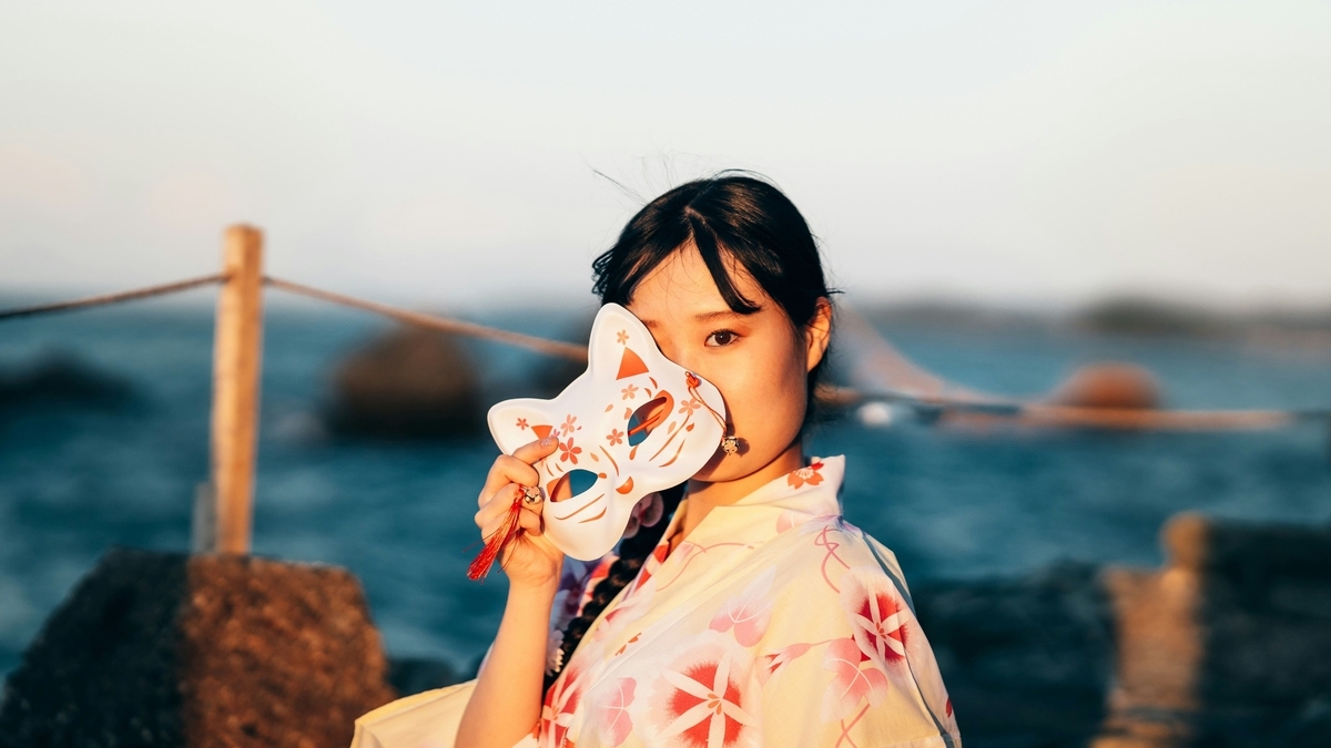 女性がキツネのお面を顔の前に掲げ、海辺の岩場に立っている。女性はピンク色の花の模様が描かれた着物を着ており、背景の海と夕日がぼけている