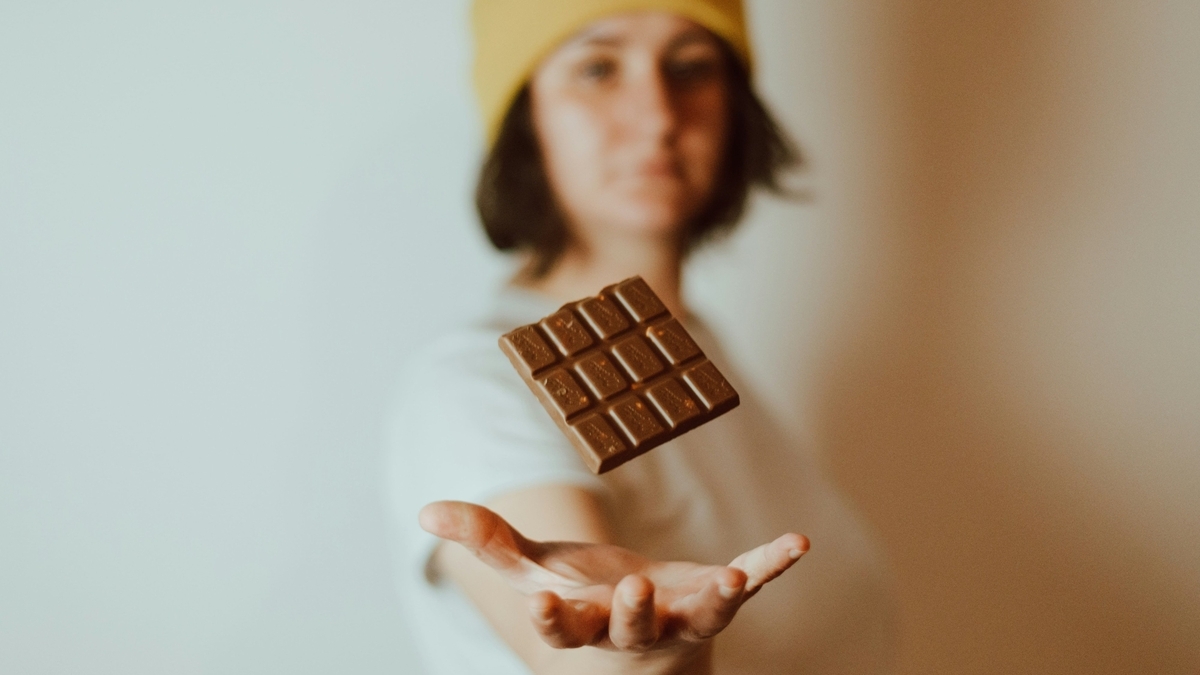 黄色いニット帽を被った人物が、掌の上に一口サイズの板チョコレートを浮かせて持っている様子。背景は白くぼかされており、チョコレートの表面の質感が強調されている