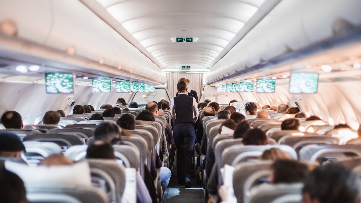 多くの乗客が座る飛行機のエコノミークラスの機内。中央の通路を紺色の制服を着た客室乗務員が歩いており、座席上部のモニターには機内エンターテインメントの映像が映し出されています