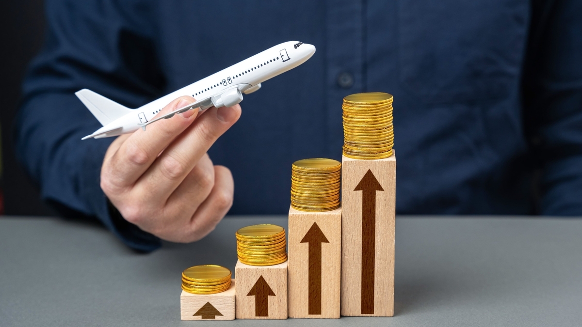 青いシャツを着た人物が模型の飛行機を手に持ち、右側には木製のブロックが階段状に積まれています。各ブロックの上には金貨が置かれ、側面には上昇を示す上向きの矢印が描かれています