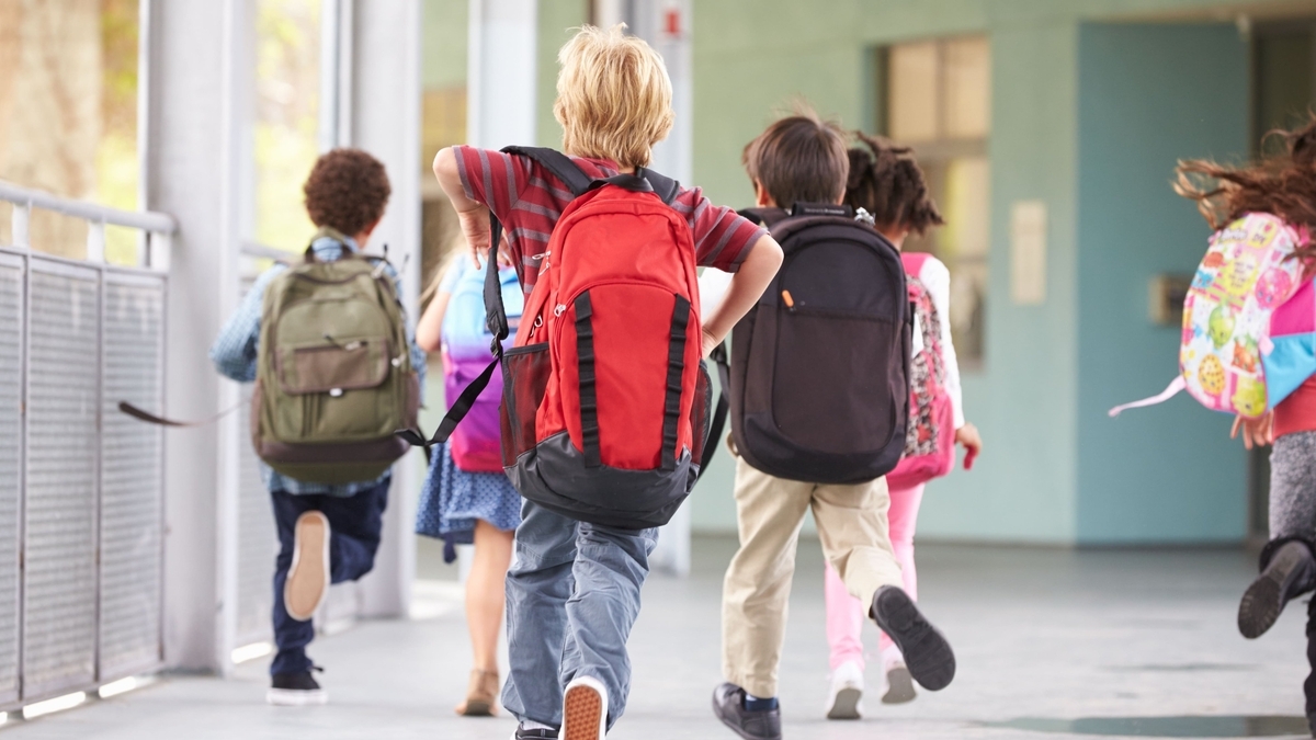 色とりどりのリュックサックを背負った小学生たちが、学校の廊下を元気に走っていく後ろ姿。手前には赤いリュックを背負った金髪の男の子が写り、開放的な校舎の様子が伺えます