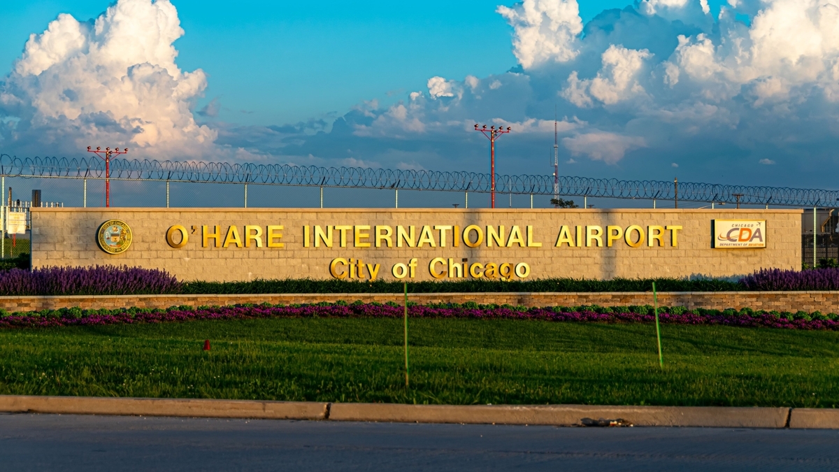 夕暮れ時のオヘア国際空港入口。石造りの壁に金色の文字で「O'HARE INTERNATIONAL AIRPORT」「City of Chicago」と刻まれ、両脇にCDAロゴとシカゴ市章がある。手前には紫の花壇と芝生がある
