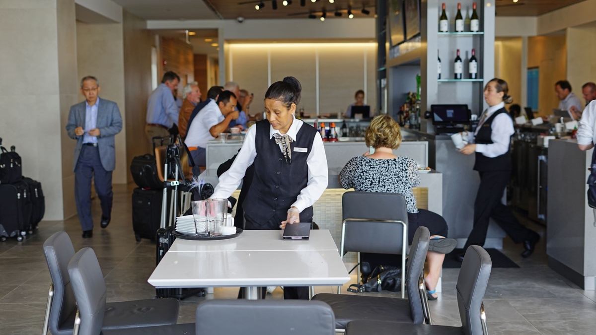 シカゴ・オヘア国際空港にある現代的な空港ラウンジ。女性スタッフが料理の載ったトレイを持って、白いテーブルに近づく様子。背景にはバーカウンターや、荷物を持つ複数の旅行者の姿が写っている