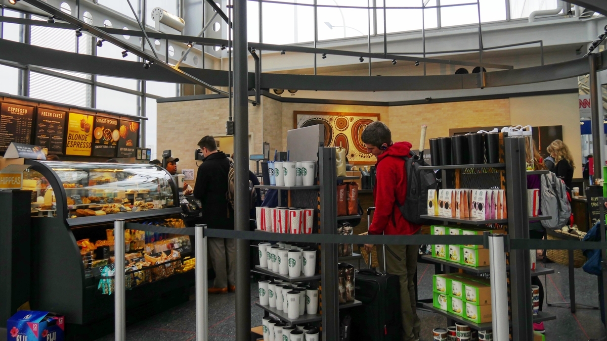 オヘア空港内のスターバックス。ガラスのショーケースには軽食やケーキが並び、その横の棚には多くのスターバックスのマグカップが陳列されている。赤い服を着た男性客が棚の商品を見ている