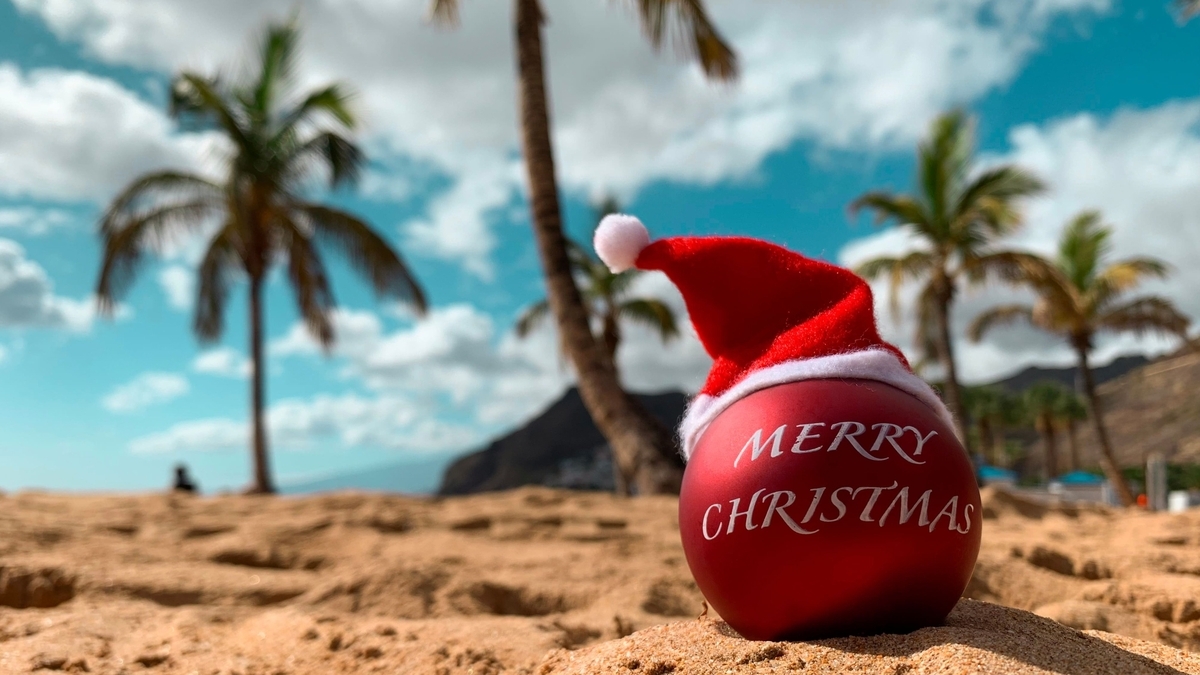 南国のビーチの砂の上に置かれた、サンタ帽子を被った赤いクリスマスオーナメント。表面には「MERRY CHRISTMAS」と白文字で書かれている。背景にはヤシの木、山、青い空と白い雲が広がっている