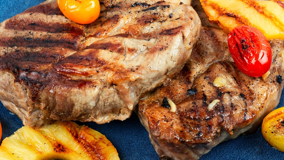 青い皿に盛り付けられた、焼き色のついたジューシーなグリルステーキ。付け合わせとして、表面を焼いた輪切りのパイナップルや、真っ赤なミニトマトが添えられており、肉の表面にはスパイスが散らされている