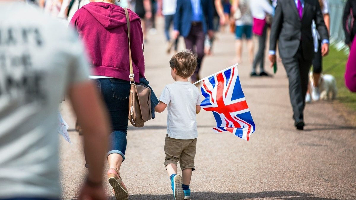 小さな子供がイギリス国旗のミニフラッグを手に持ち、紫色のパーカーを着た大人の隣を歩く様子。周囲には多くの人々が行き交い、奥にはスーツ姿の男性や白い犬を連れた人の姿も見えます
