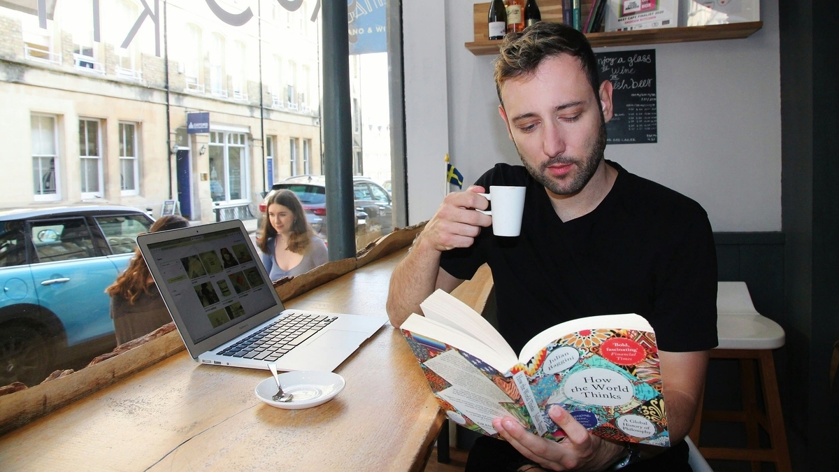 カフェの窓際にある木製カウンター席で、男性が本を読みながらコーヒーを飲む様子。手元には開かれた本とノートパソコンがあり、窓の外には街並みや通りを走る青い車、歩行者の姿が映っています