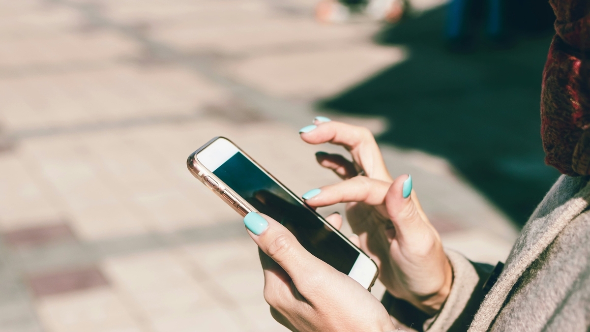 日差しが降り注ぐ屋外で、水色のネイルをした女性が金色のスマートフォンを両手で持ち、指先で画面を操作している様子