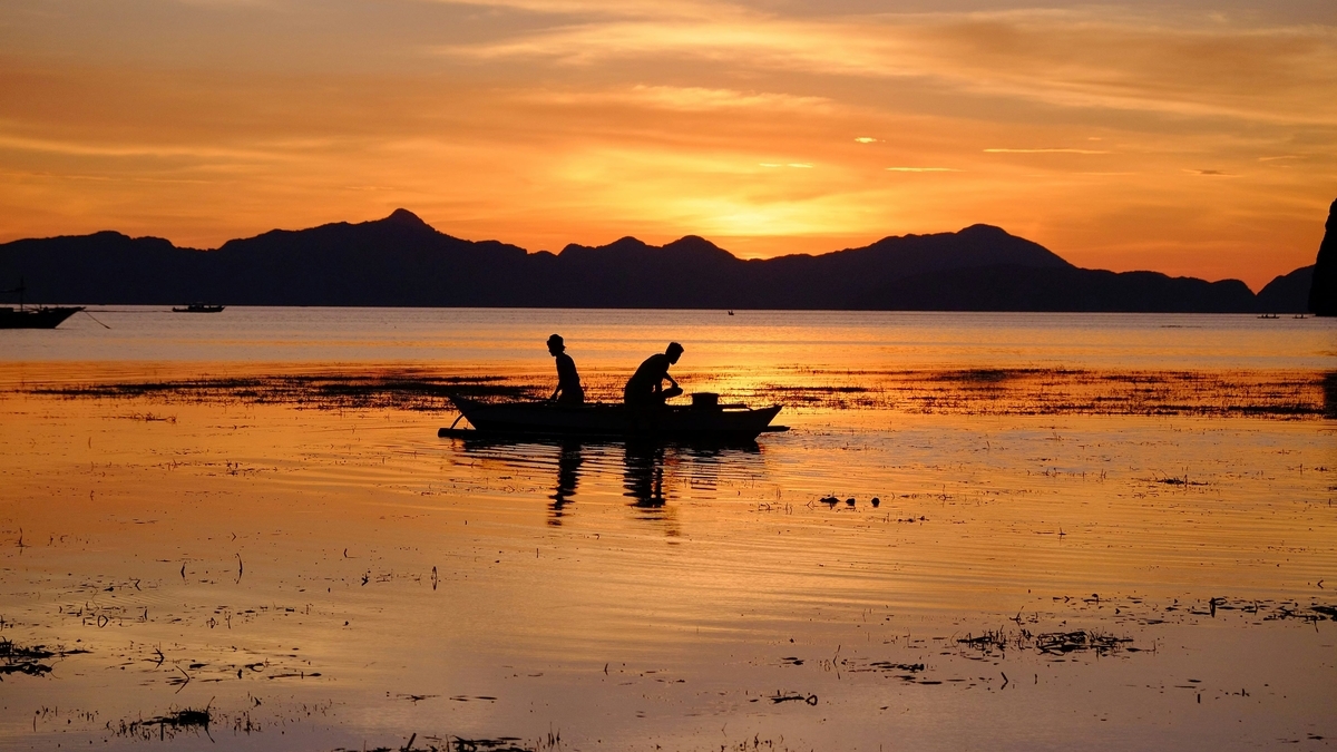 オレンジ色に染まる夕暮れ時の海で、山々を背景に小さな舟の上で作業をする二人の人物のシルエット。水面には夕日の光が反射している