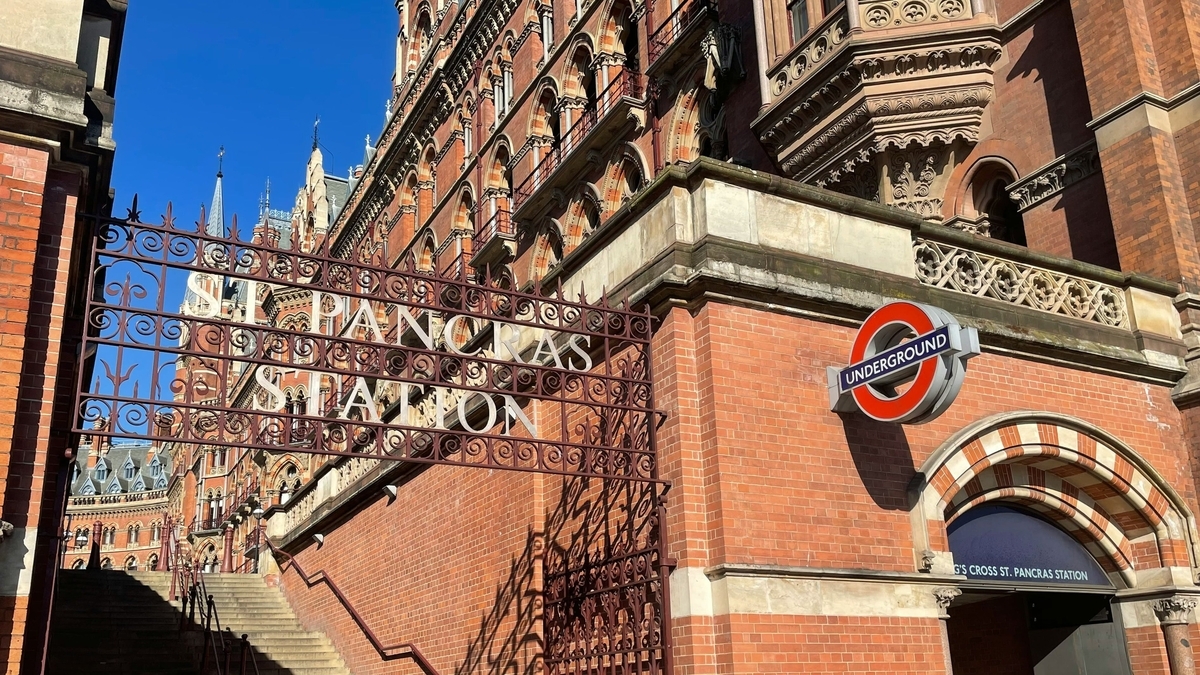 ロンドンのセント・パンクラス駅のレンガ造りの外壁と、アイアンゲートに刻まれた駅名の文字。右手には地下鉄「UNDERGROUND」の赤いロゴマークが掲げられ、青空が広がっている