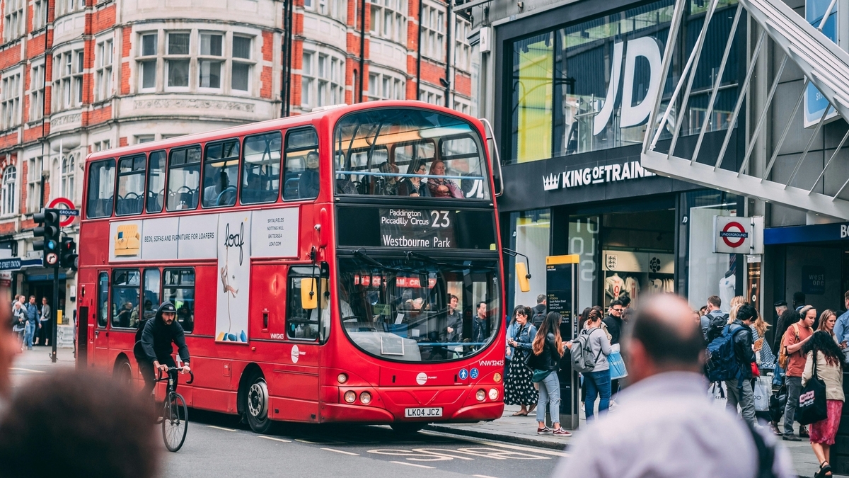 ロンドンの街角を走る赤い二階建てバス。行き先表示には「Westbourne Park」とあり、周囲には多くの歩行者やショップ、自転車に乗る人物が行き交っている