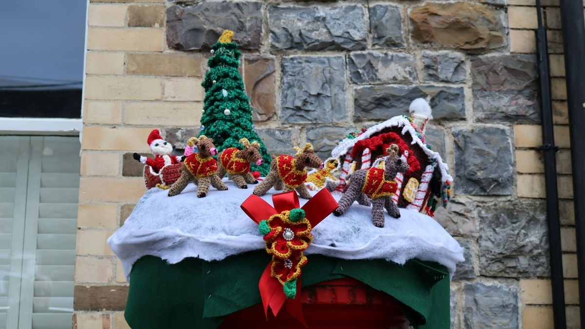 レンガの壁を背景に、雪に見立てた白い綿の上に置かれた手編みのクリスマス飾り。サンタクロースやトナカイ、ツリー、小さな家が並び、中央には赤いリボンと花の編み物が飾られている