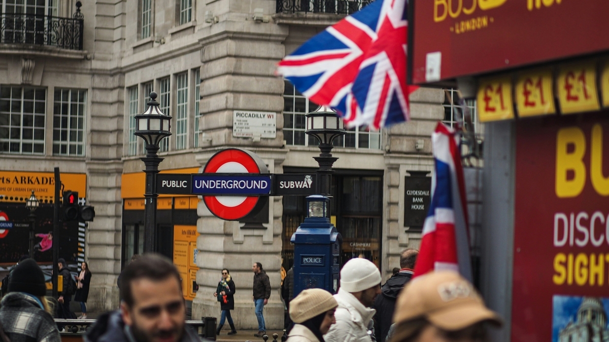 ロンドンのピカデリー・サーカス付近の街並み。赤と白の地下鉄ロゴマーク「アンダーグラウンド」の看板が中央にあり、背景にはイギリス国旗や行き交う人々、歴史を感じさせる石造りの建物が写っている