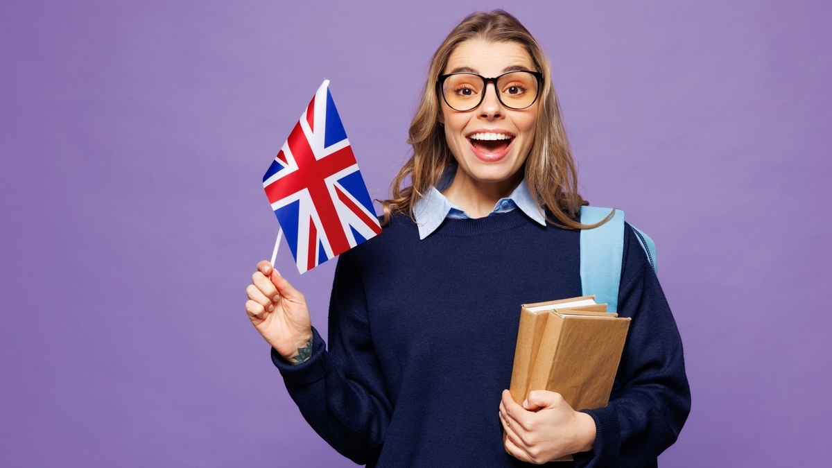 紫色の背景の前で、眼鏡をかけ本を抱えた笑顔の女性が、右手で小さなイギリスの国旗を掲げている姿