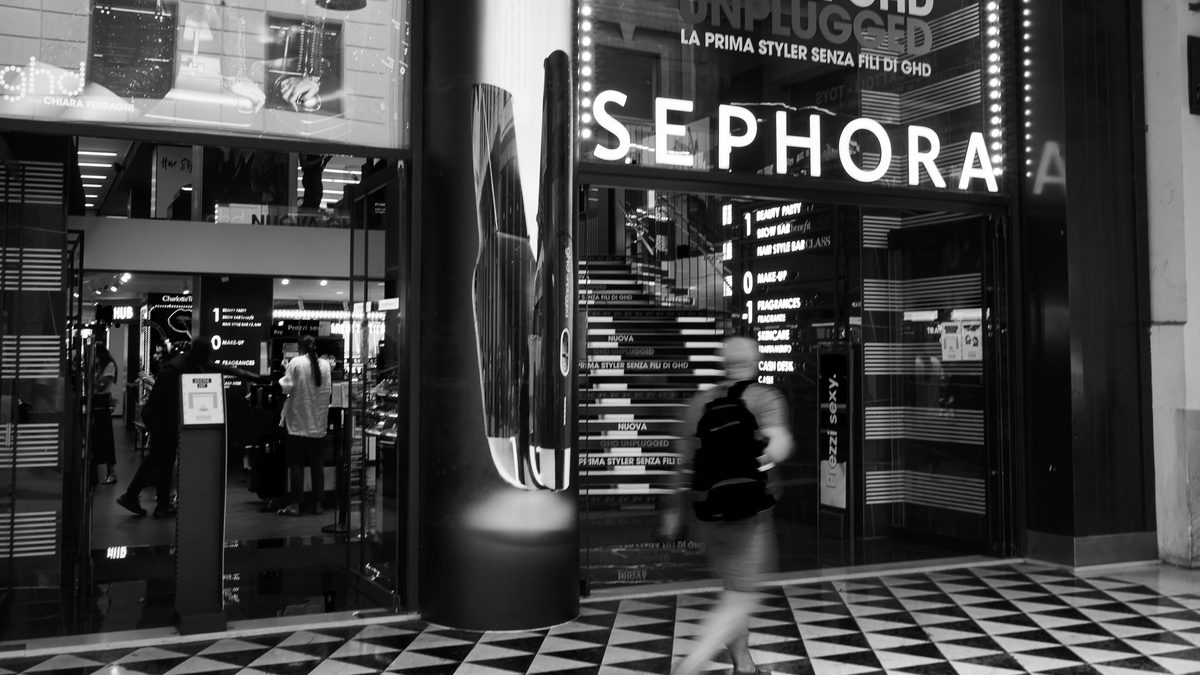 モノクロ写真で捉えられたセフォラの店舗入り口。ガラス扉の上には大きなSEPHORAのロゴがあり、店内には商品棚や階段、フロア案内が見えます。歩道にはリュックを背負った通行人の姿が写っています