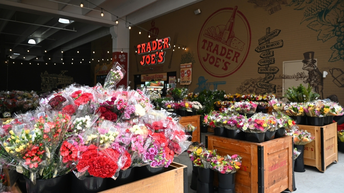 トレーダー・ジョーズの店内に並ぶ色鮮やかな切り花の束。手前には赤いカーネーションが大量に配置され、壁面にはお店のロゴやイラスト、地名が書かれた標識のようなデザインが描かれています