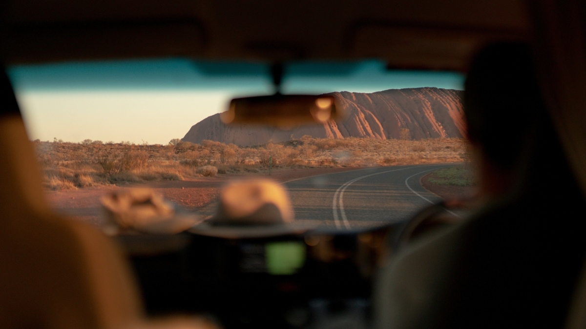 走行中の車のフロントガラス越しに眺める、夕日に照らされたウルルの風景。ダッシュボードにはハットが置かれ、前方の道路は緩やかなカーブを描きながら巨大な岩山へと続いています。車内から捉えた旅のワンシーンです