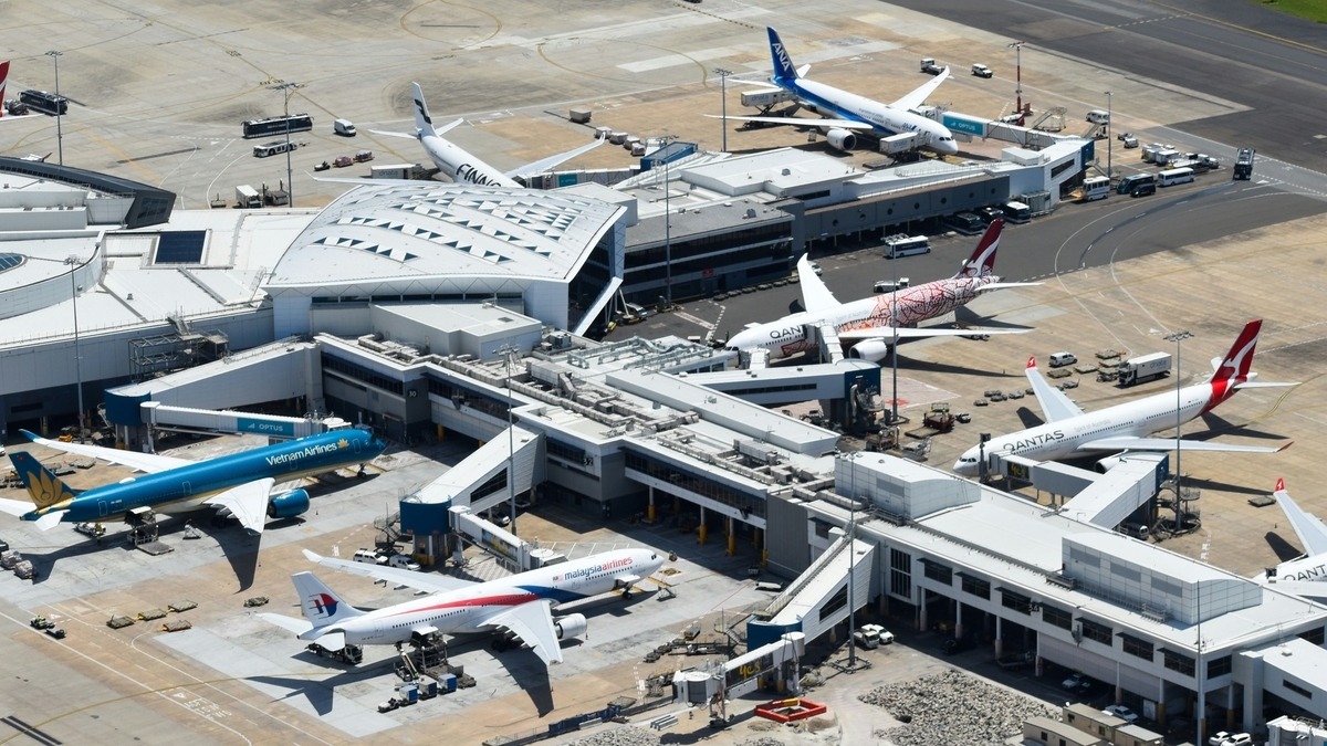 晴天の空の下に広がる空港の全景。マレーシア航空やカンタス航空、ベトナム航空などの機体が搭乗ゲートに並び、白い屋根のターミナルビルや、滑走路に待機する地上作業車が俯瞰で捉えられています