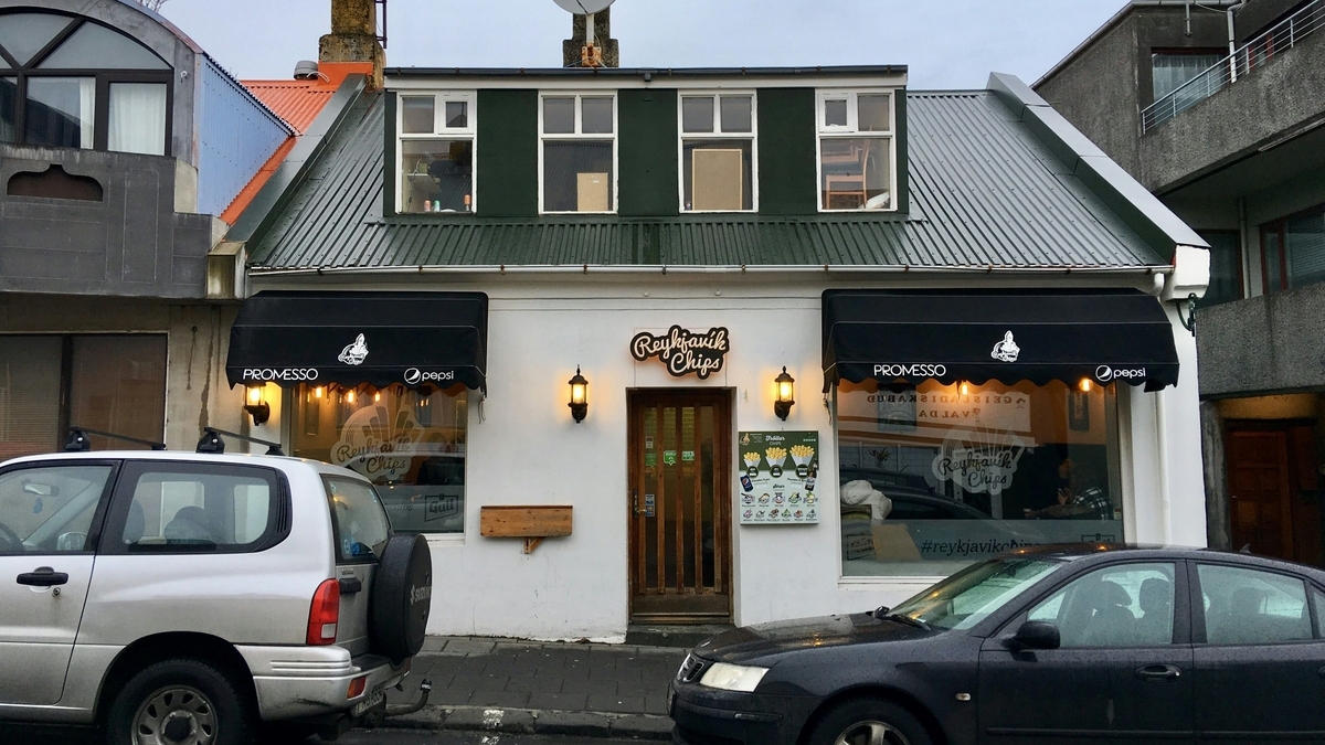 「Reykjavik Chips」と書かれた看板を掲げる、白い外壁と緑色の屋根の平屋。入り口の両脇には黒いオーニングが張り出し、暖かい光の街灯が灯っています。建物の前にはシルバーと黒の乗用車が停車しています
