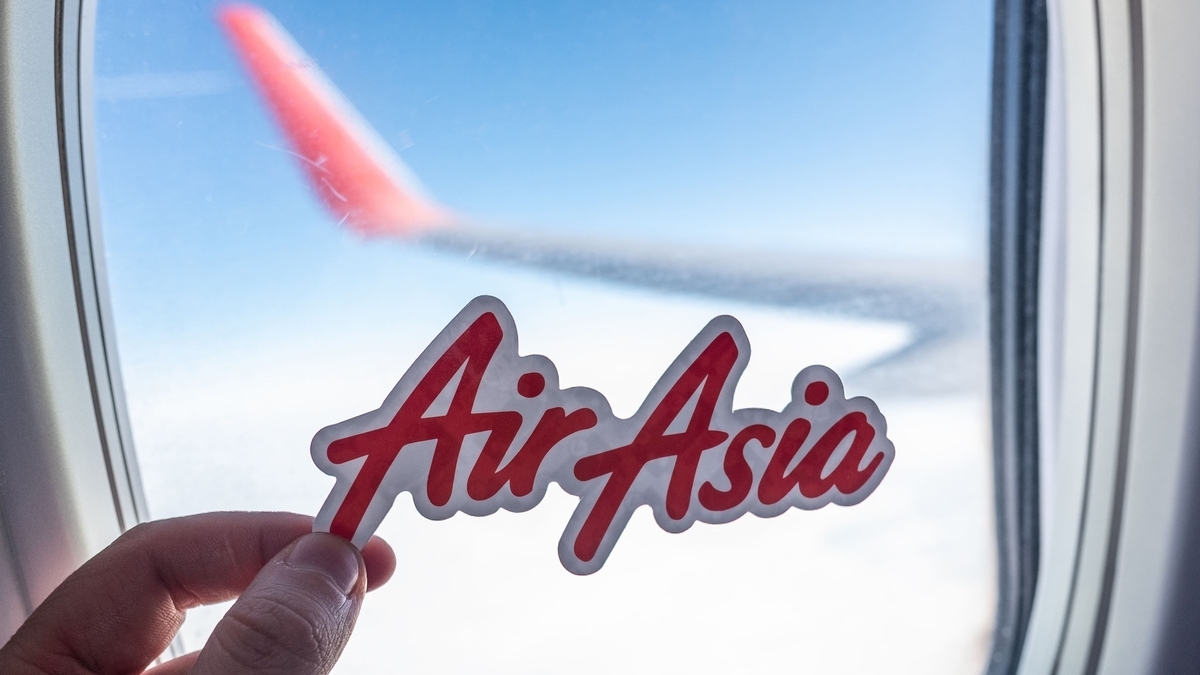 飛行機の窓越しに、エアアジアの赤いロゴステッカーを指で持っている様子。背景には青い空と雲、そして赤く塗装された機体のウィングレットがぼやけて写っています