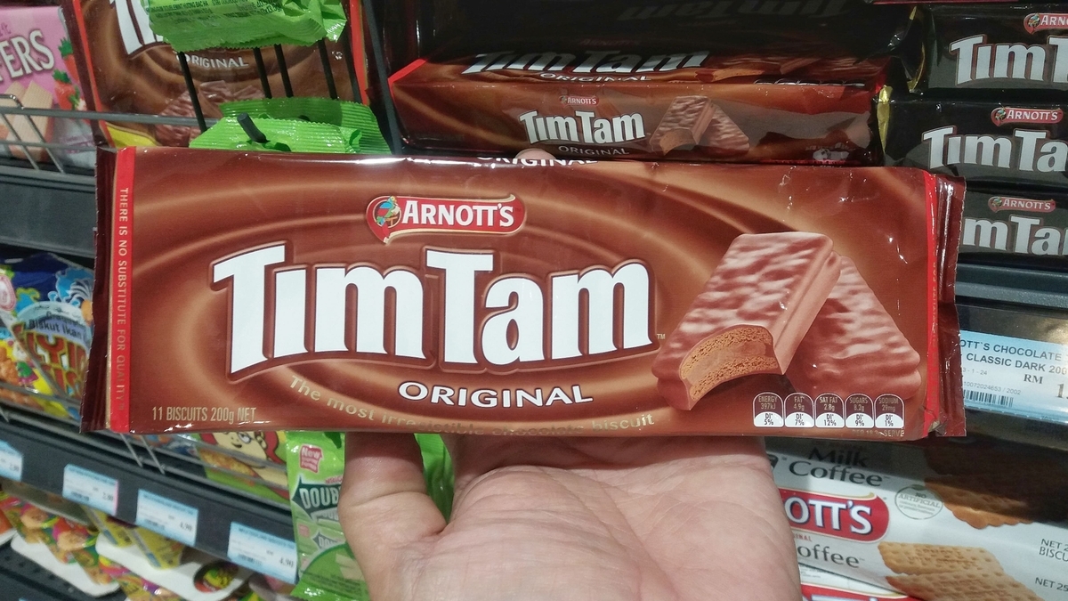 「Tim Tam ORIGINAL」と書かれた茶色のパッケージを、人の手が持ち上げている様子。パッケージには、半分に割られたチョコレートビスケットの断面写真が大きくプリントされています。