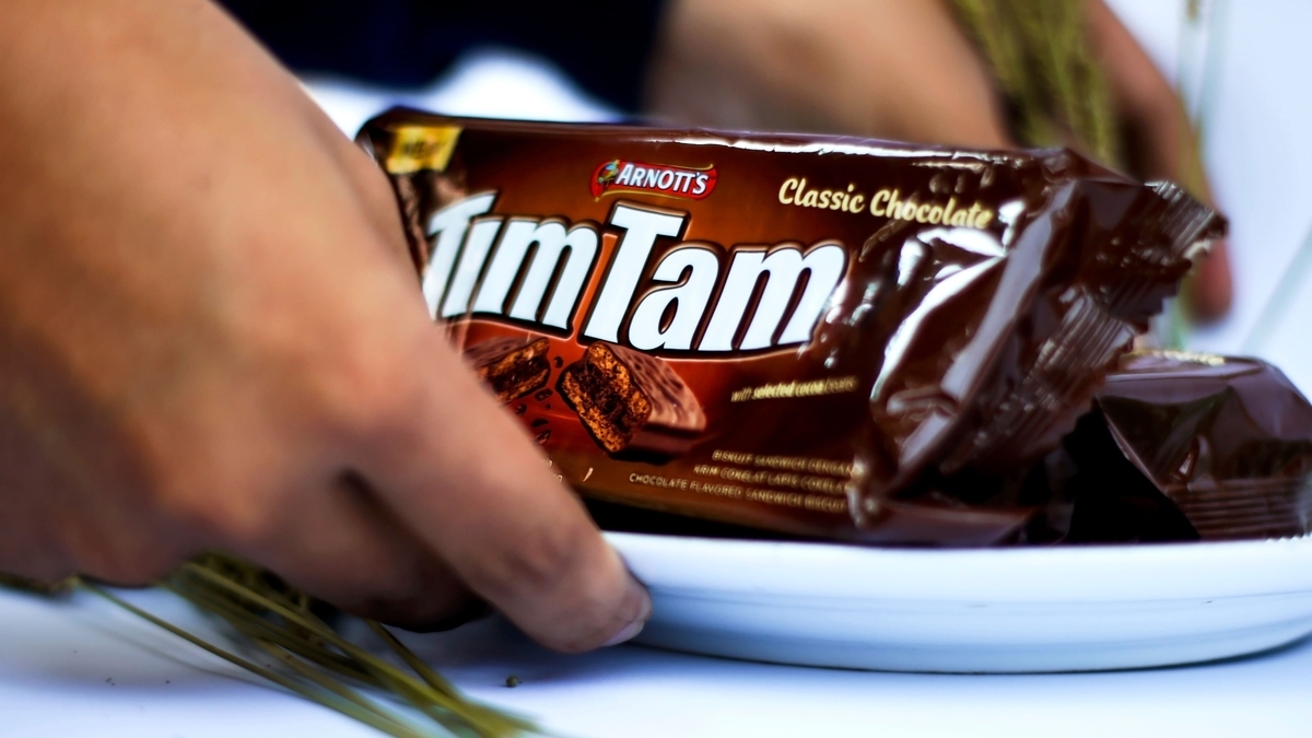 「Tim Tam Classic Chocolate」のパッケージを、白い皿の上に乗せて両手で支えている様子。背景は白く、手前には植物の茎のようなものが添えられています