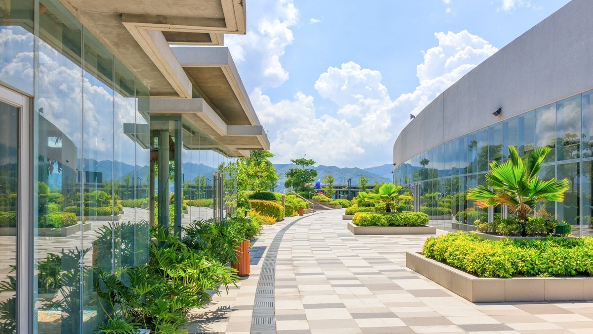 青空の下に広がる屋上庭園の風景。タイル敷きの緩やかなカーブを描く通路の両脇には、ガラス張りの建物と美しく整えられた緑の植栽が並んでいます。遠くには山々が薄く見え、開放感のある屋外空間が広がっています